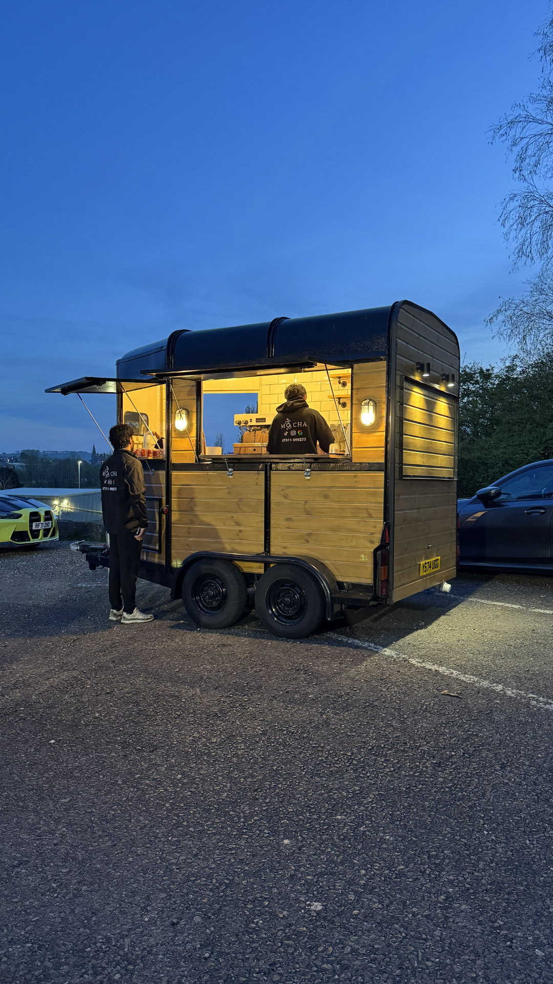 A mobile food truck serving coffee at dusk, with two people inside and one person waiting outside, parked in a lot with cars around.