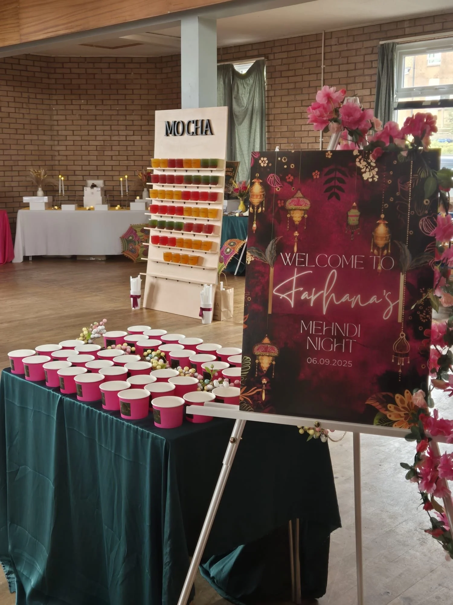 Event setup with a sign welcoming to Kahanna's Mehndi Night, date 06.09.2025, and tables with pink and white containers, decorated with flowers and a mochi stand with colorful dessert cups.