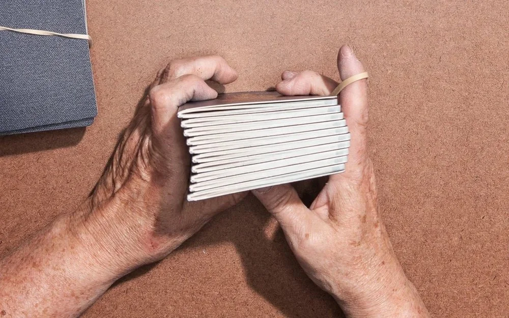 Close-up of elderly person's hands holding a small stack of white notebooks with a rubber band around them on a brown surface.