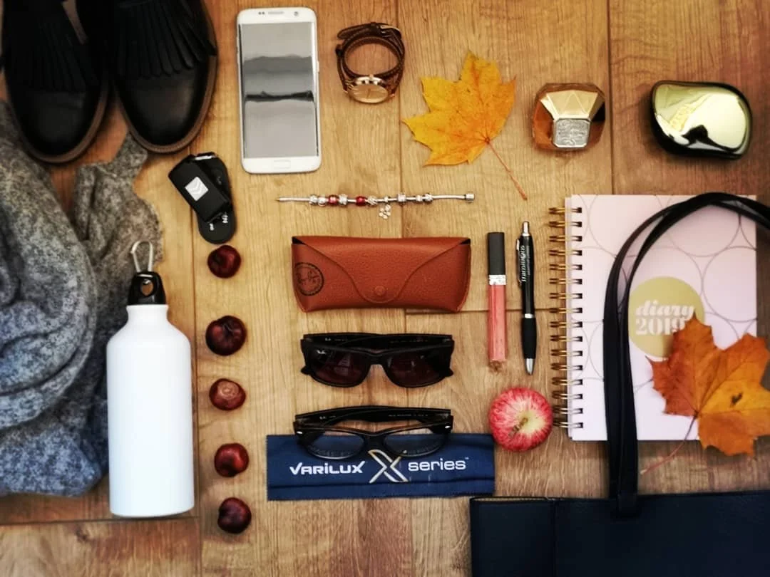 Flat lay of personal belongings on a wooden surface, including shoes, a smartphone, jewelry, sunglasses, a water bottle, snacks, a diary, and autumn leaves.