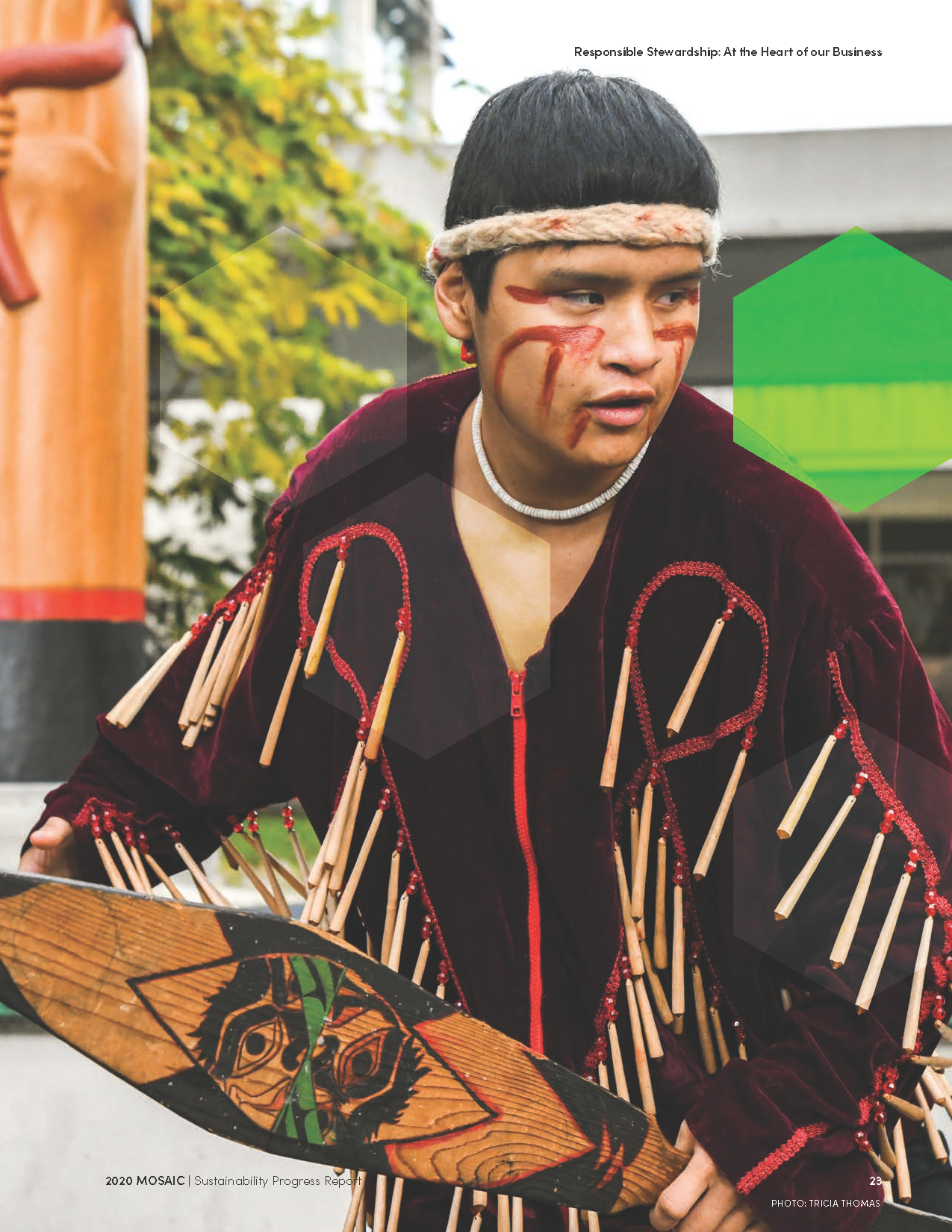 Person dressed in culturally traditional attire, holding a painted wooden shield, with face paint and a headband, outdoors near greenery and a shrine or totem pole.