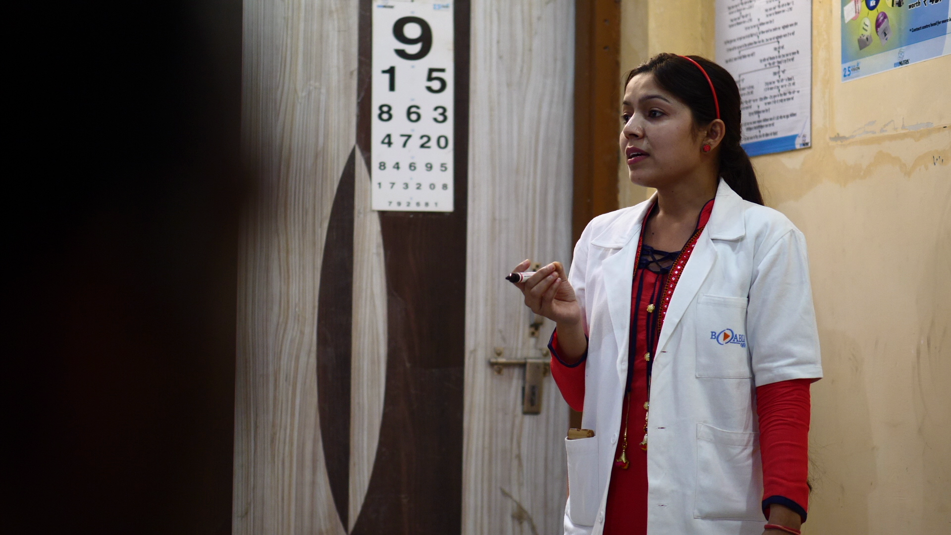 A woman in a white medical coat with a logo on the chest, holding a marker, standing in a room with a vision chart on the wall behind her.