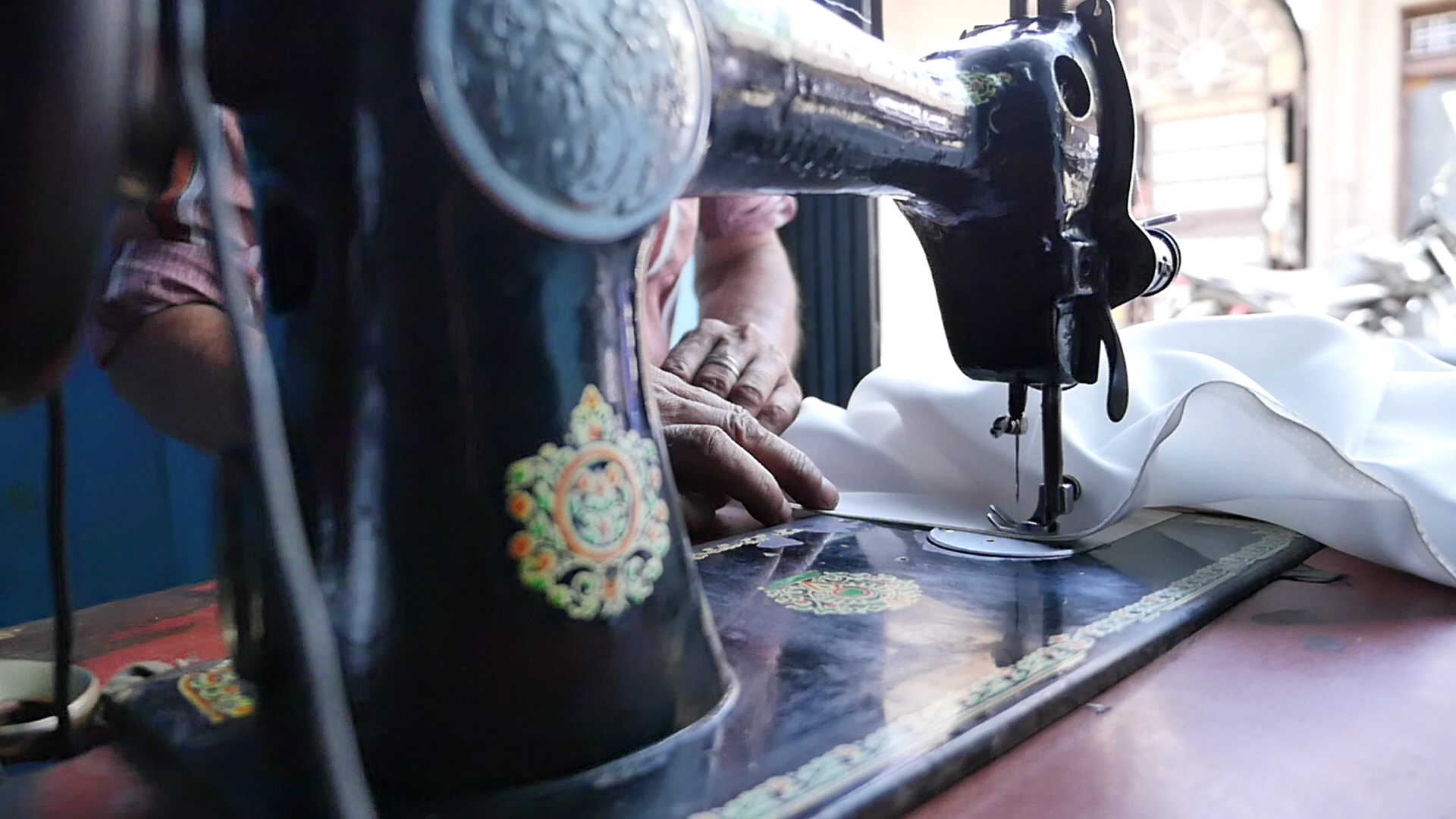Close-up view of a vintage sewing machine with black body and colorful floral designs, a person sewing white fabric with their hands visible in the background.