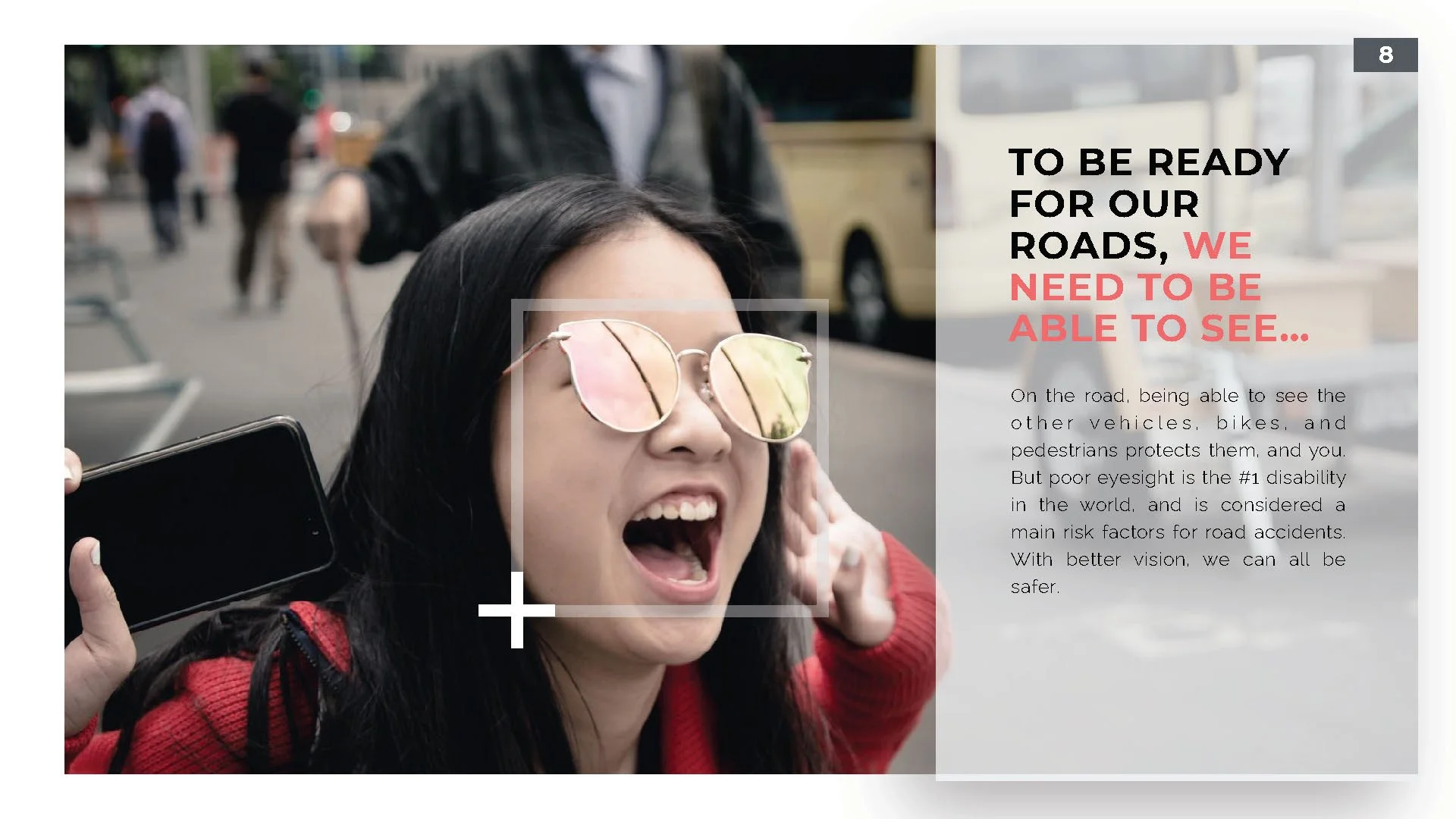 A woman wearing glasses and a red jacket covering her ear, holding a smartphone, in an outdoor urban setting.