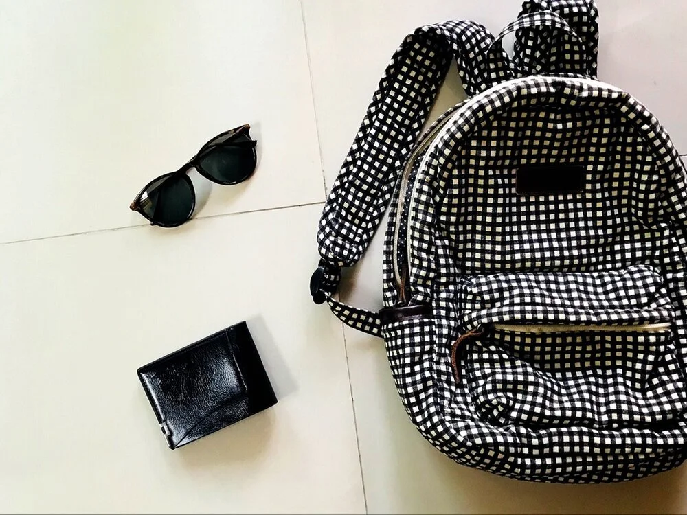 Black sunglasses, black wallet, and a black and white checkered backpack on a white tiled floor.
