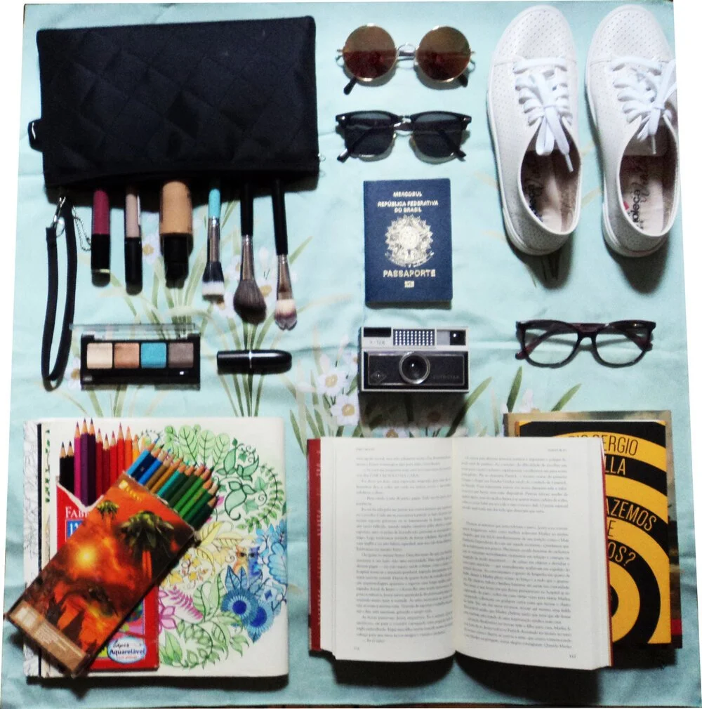 Travel essentials including makeup brushes, sunglasses, white sneakers, eyeglasses, a passport, a camera, books, colored pencils, and a box of colored pencils on a light green floral surface.