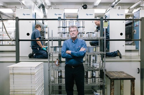 A man standing with arms crossed in front of industrial machinery, with two workers in the background operating equipment in a manufacturing or industrial setting.