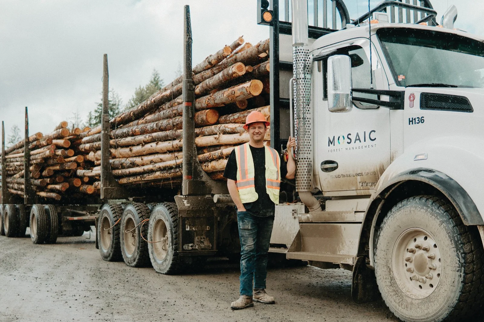 A man in a safety vest and hard hat standing next to a logging truck loaded with logs on a dirt road in a forested area.
