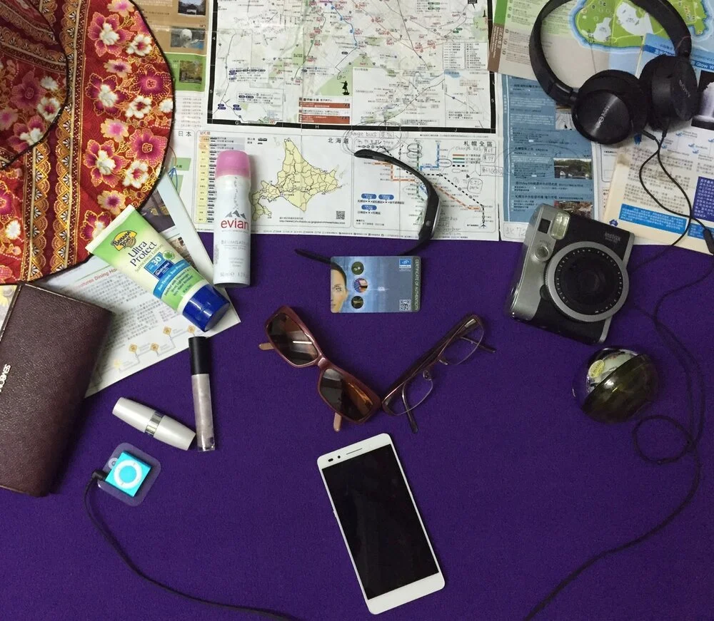 A traveler's cluttered table with maps, a camera, sunglasses, a smartphone, headphones, sunscreen, and a lipstick on a purple surface.