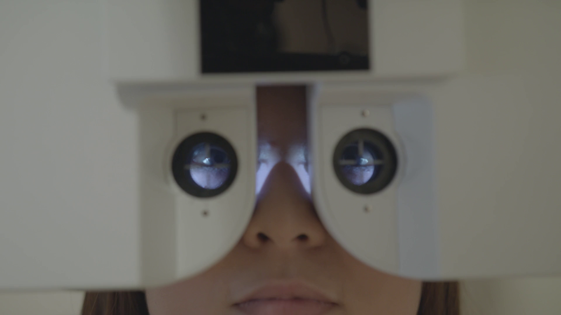 Close-up of a person's face looking through an eye examination device, with the device's circles and mirrors visible around the person's eyes.