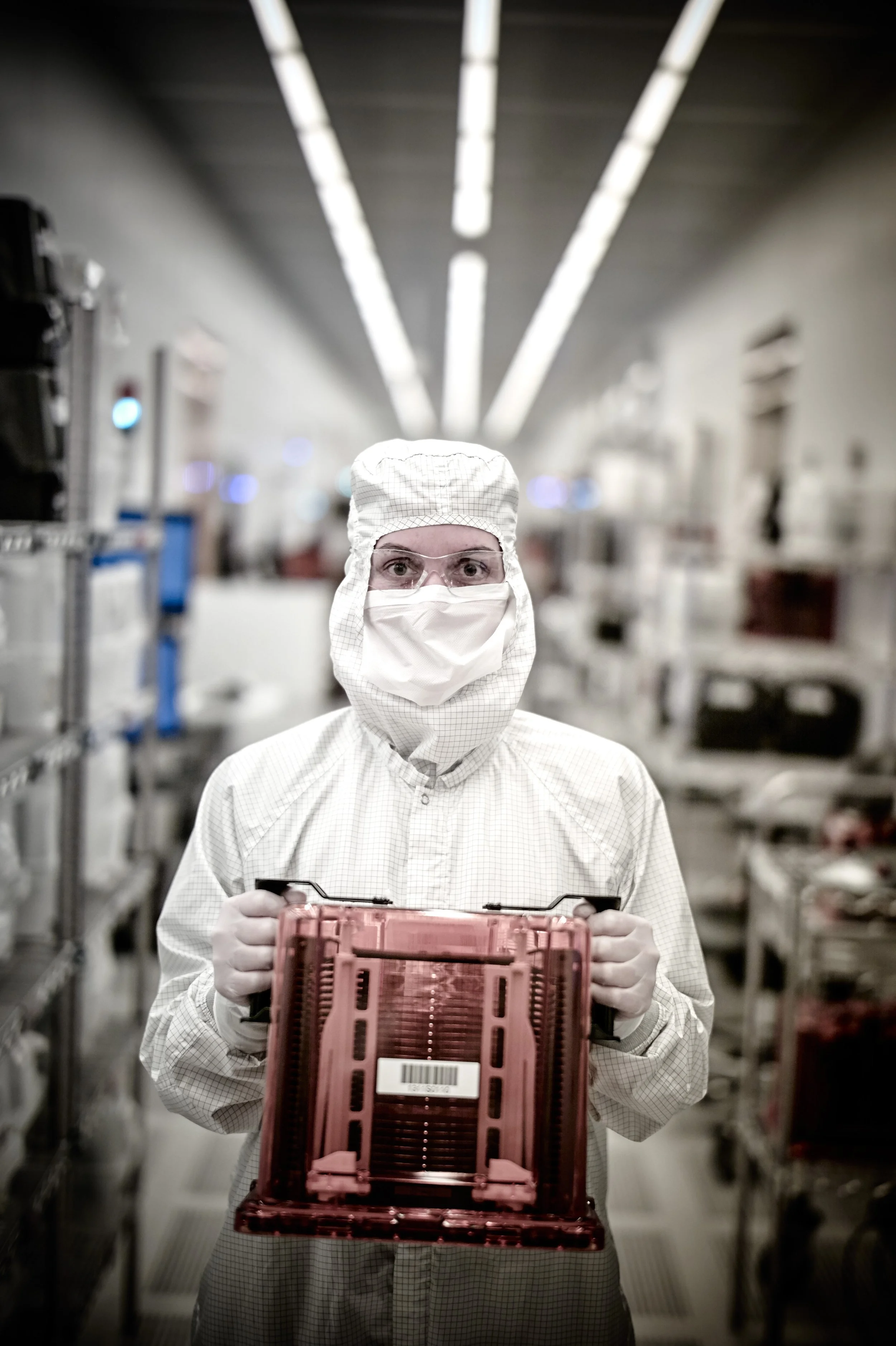 A worker in protective clothing, mask, and glasses holding a pink plastic tray in a processing or manufacturing facility.
