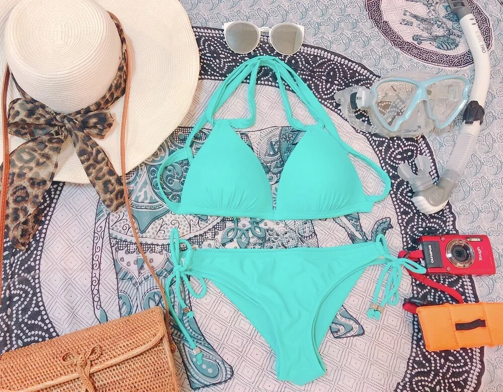 Beach accessories including a wide-brimmed hat with a leopard-print scarf, a pair of sunglasses, a turquoise bikini, snorkeling gear, a red disposable camera, an orange waterproof case, and a woven purse on a patterned beach towel.