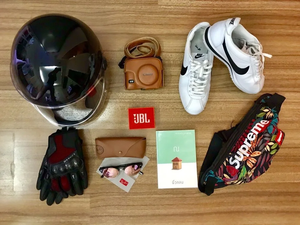 Various personal items laid out on a wooden floor, including a black motorcycle helmet, brown camera case, Nike sneakers with swoosh logo, Supreme-branded fanny pack with floral pattern, red and black gloves, sunglasses in a brown case, JBL logo, and