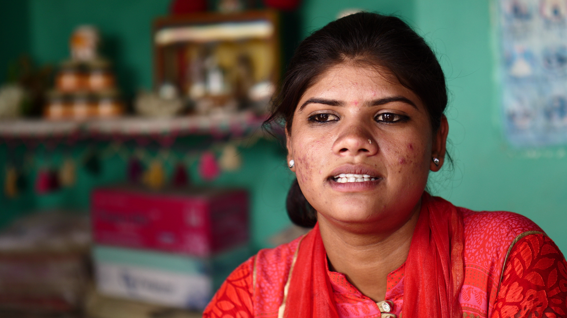 A young woman in traditional Indian clothing, with bacterial acne scars on her face, looking pensively to the side, with a blurred background of a green wall and shelves.