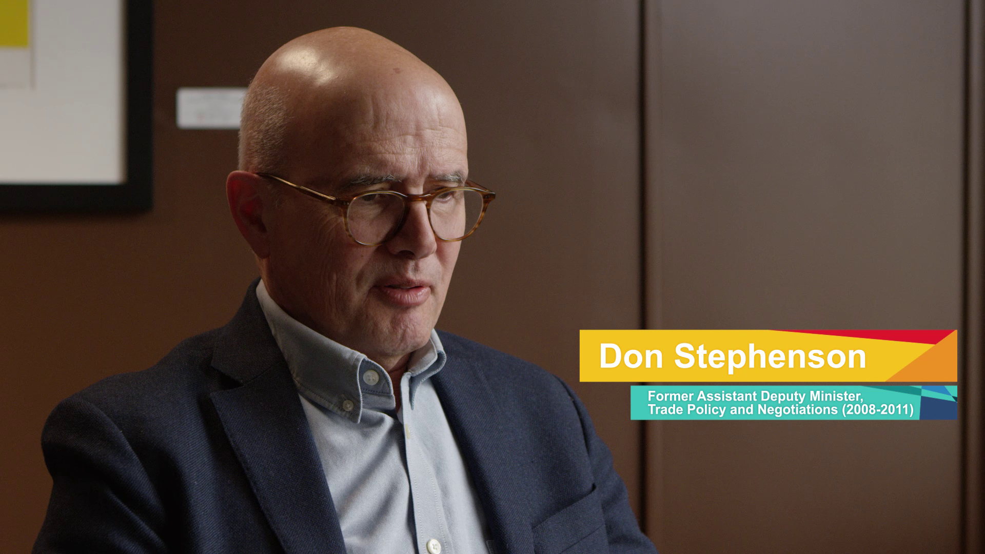 A man with glasses and a bald head, wearing a suit jacket and shirt, looking downward in an indoor setting with a brown wall background and a framed picture.