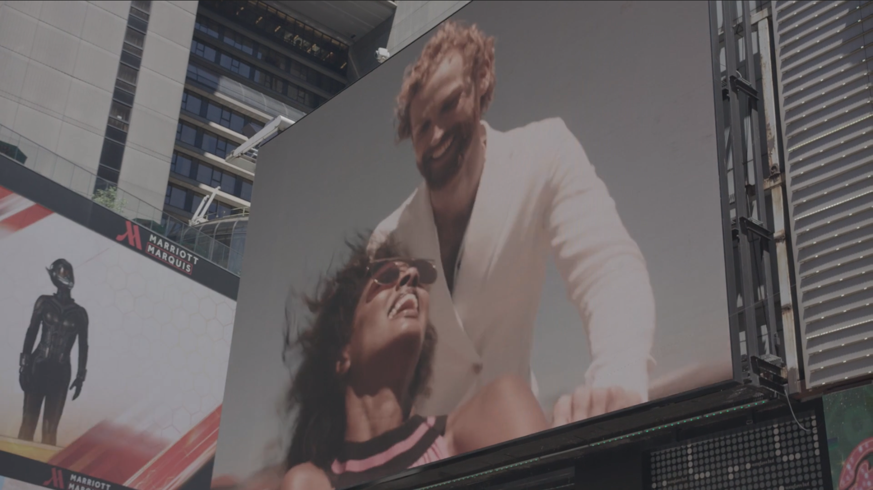 Large billboard showing a man and a woman smiling and laughing, the man wearing a white suit and the woman wearing sunglasses, against a plain background.