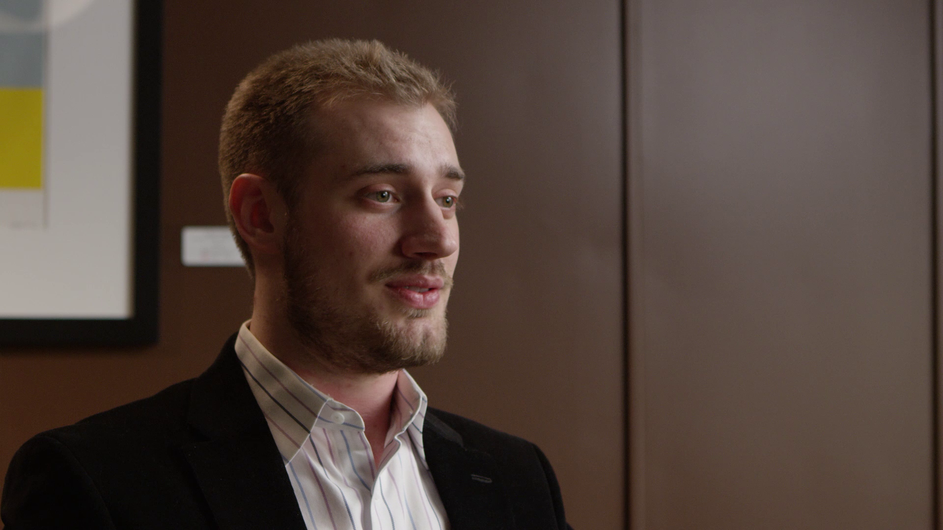 A young man with light brown hair and a beard, wearing a dark blazer and a light striped shirt, standing indoors against a brown wall with framed art.