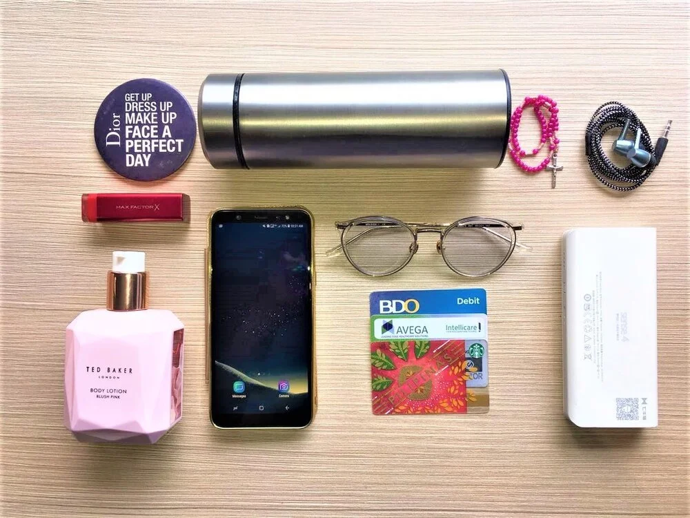 A flat lay of personal items on a wooden surface, including a round Dior makeup compact, a stainless steel travel mug, pink and black beaded bracelets, a small red Max Factor lip balm, a pink Ted Baker body lotion, a smartphone, a pair of round eyegl