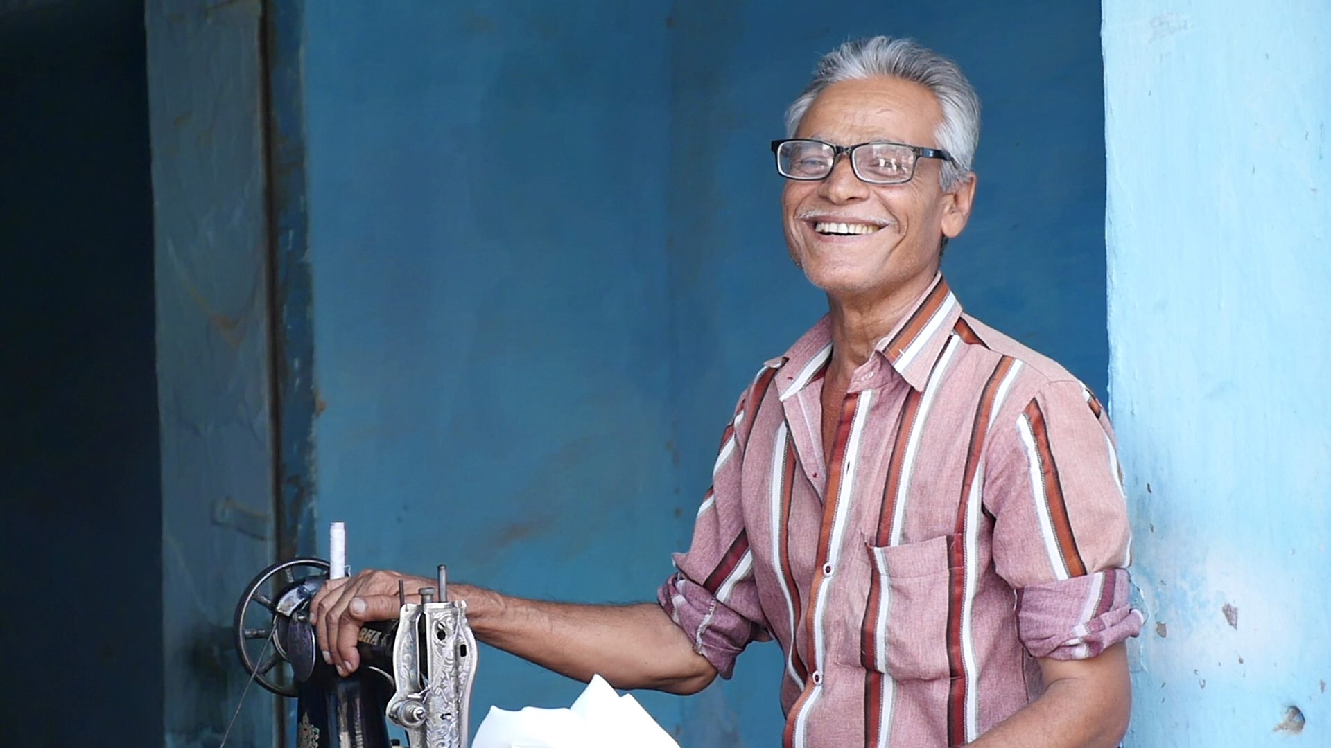 Smiling elderly man with glasses in a short-sleeved, striped shirt standing beside a sewing machine against a blue wall.