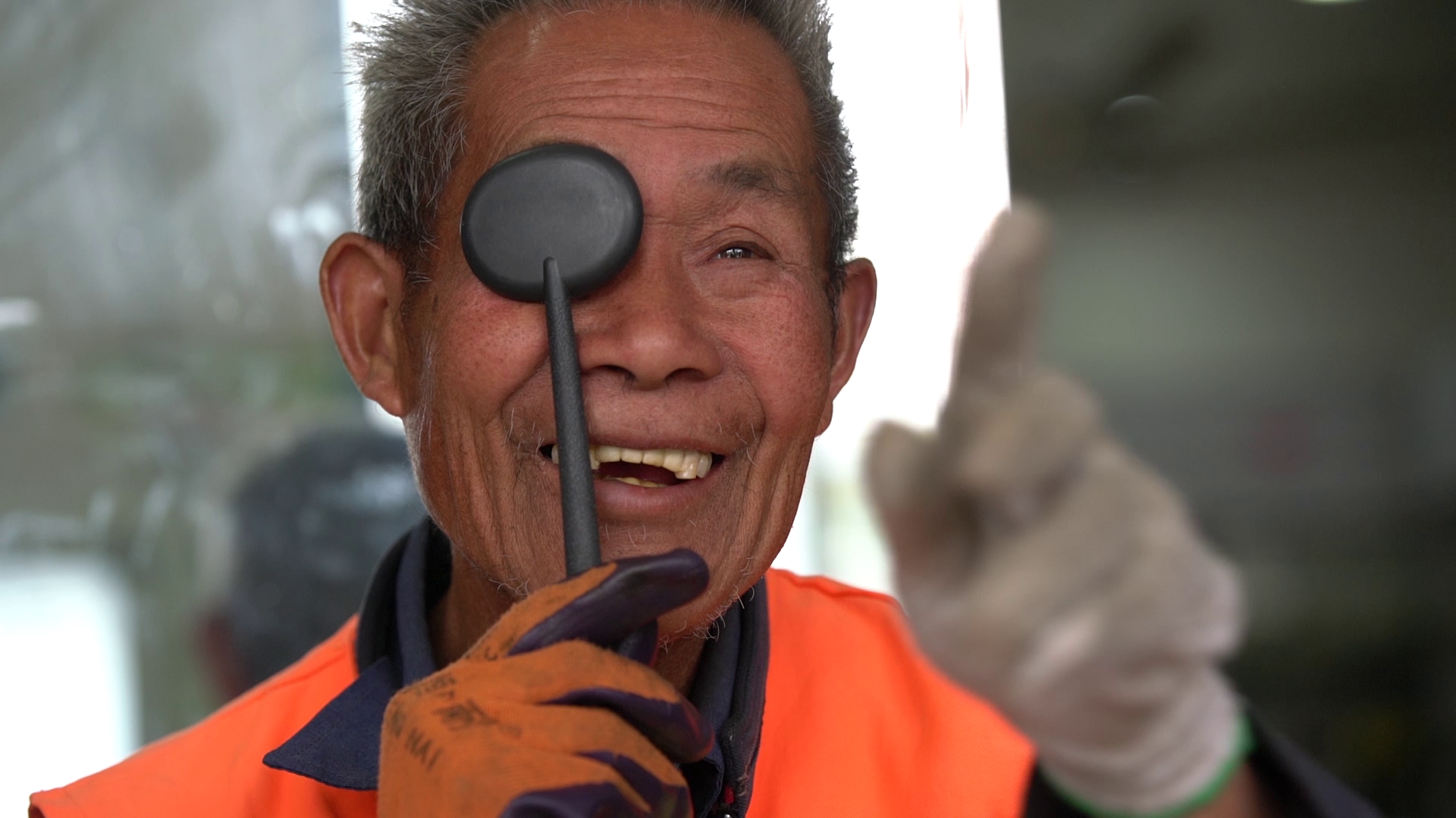 A smiling elderly man in an orange safety vest holds a black magnetic pick-up tool near his eye, pointing it at the camera. He is wearing gloves and giving a thumbs-up.