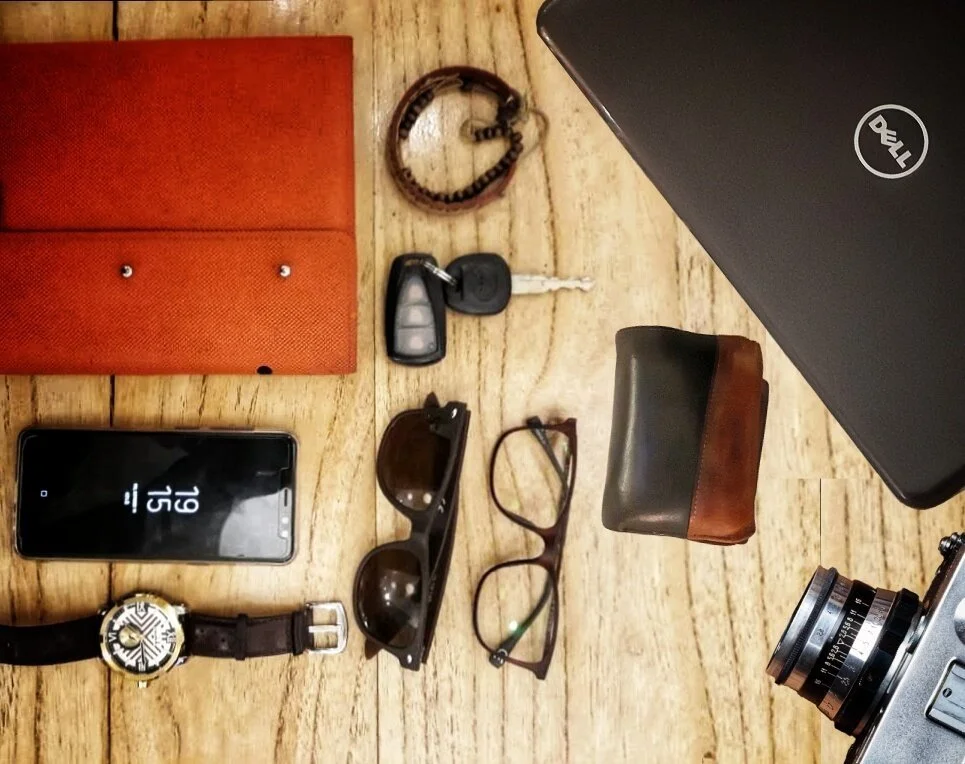 Flat lay of personal items on a wooden surface, including a red notebook, a phone displaying the time 3:15, sunglasses, a watch, a pair of glasses, a small brown and black pouch, a leather bracelet, a set of car keys, a red and black Dell laptop, and