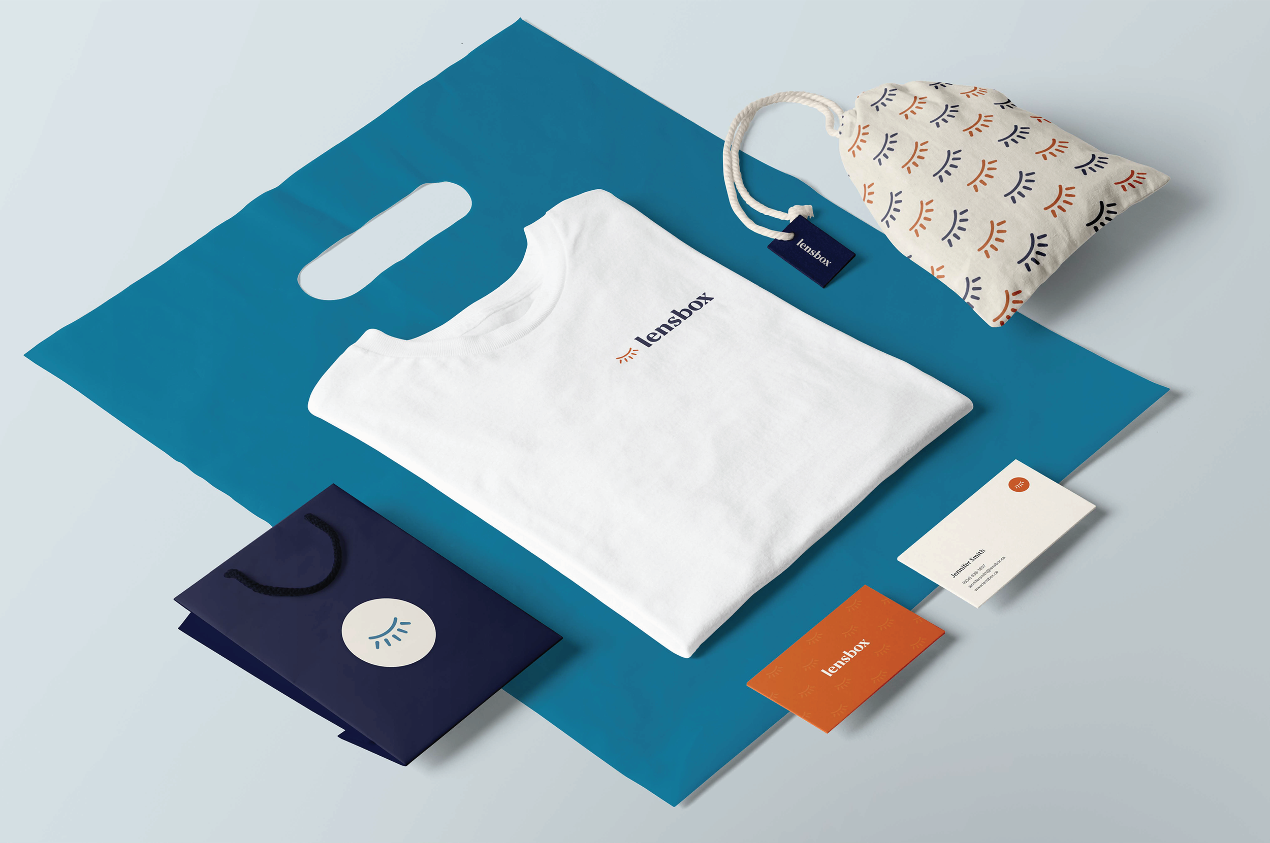 Branding materials including a white T-shirt with the Lensbox logo, a navy gift bag with a lens icon, a patterned fabric pouch, a blue shopping bag, and various business cards and tags, all arranged on a blue surface.