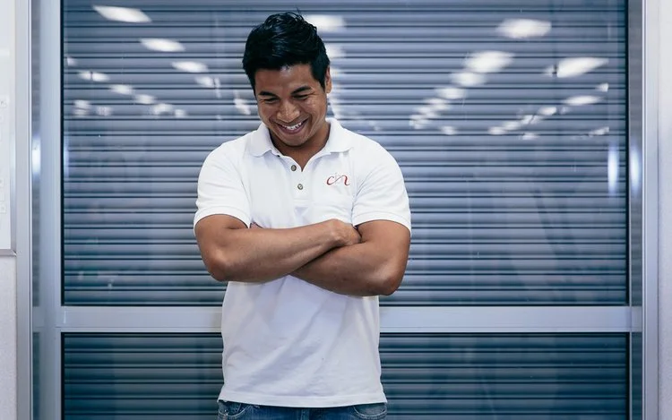 A man standing in front of a metallic, slatted wall, wearing a white polo shirt with a logo, smiling with arms crossed.