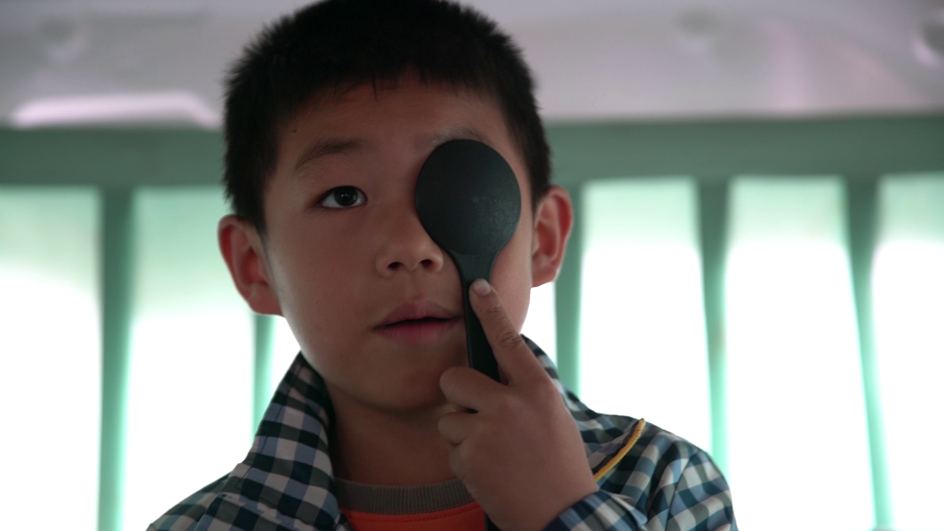 A young boy looking at the camera while holding a black spoon over his right eye.
