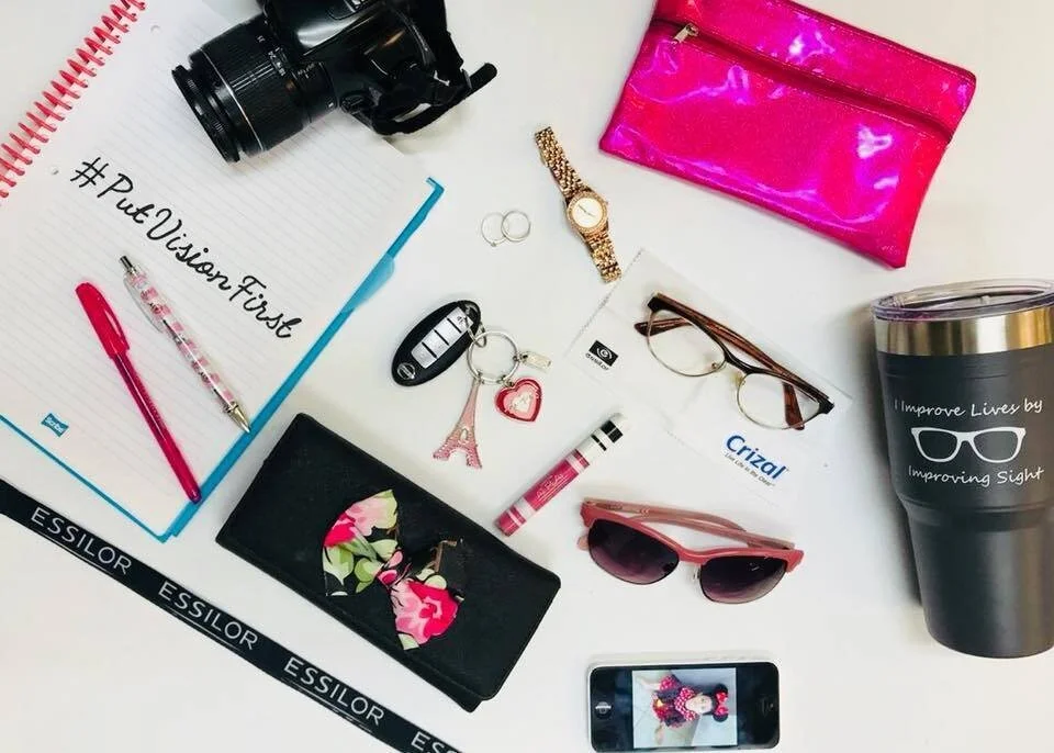 Cluttered desk with camera, pens, notebook with hashtag, wallet with floral design, sunglasses, pink glitter clutch, watch, keychain with Eiffel Tower and heart, crizal glasses, a pink crayon, a travel mug with sunglasses logo, and a smartphone showi