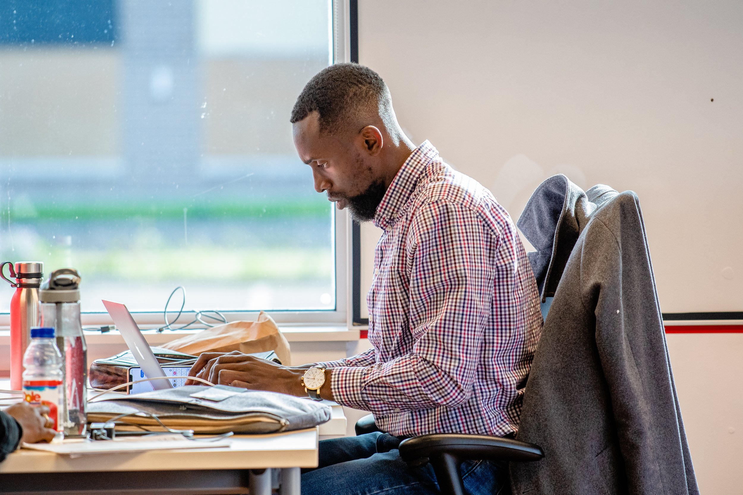 Man working on a laptop at a desk with water bottles and papers, sitting next to a window.