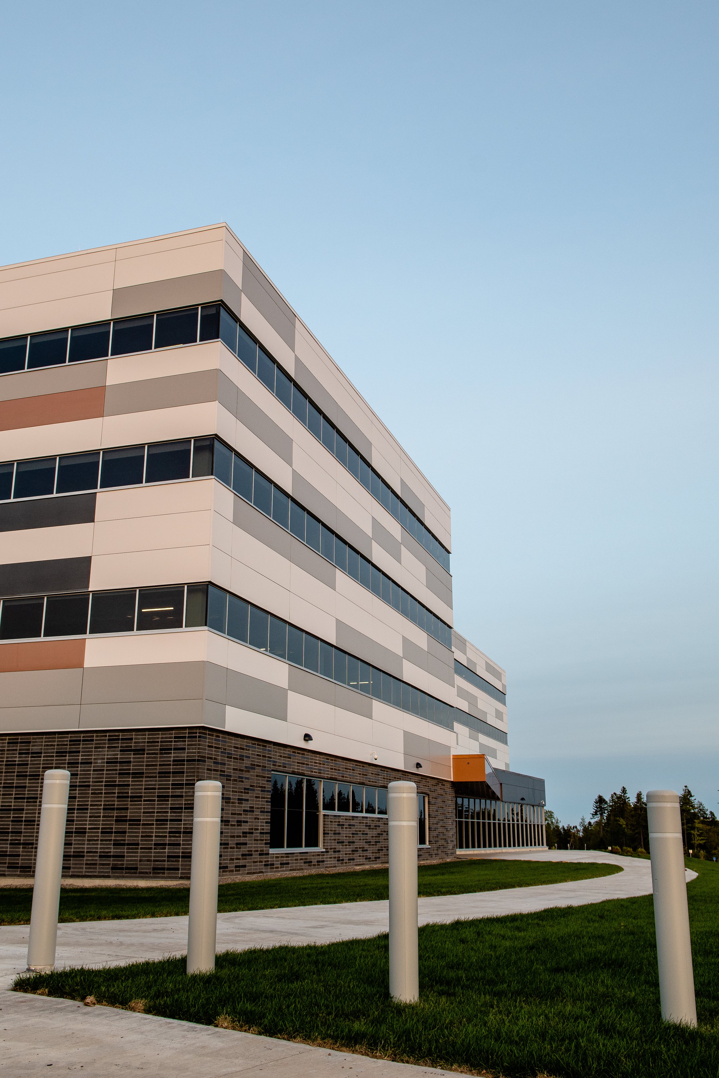 Modern multi-story building with large windows, situated next to a grassy area with a paved walkway and white posts, under a clear sky.