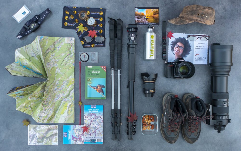 Hiking and photography gear arranged on a gray surface, including a foldable map, compass, camera with telephoto lens, hiking boots, trail notebook, water bottle, hat, sunglasses, and travel accessories.