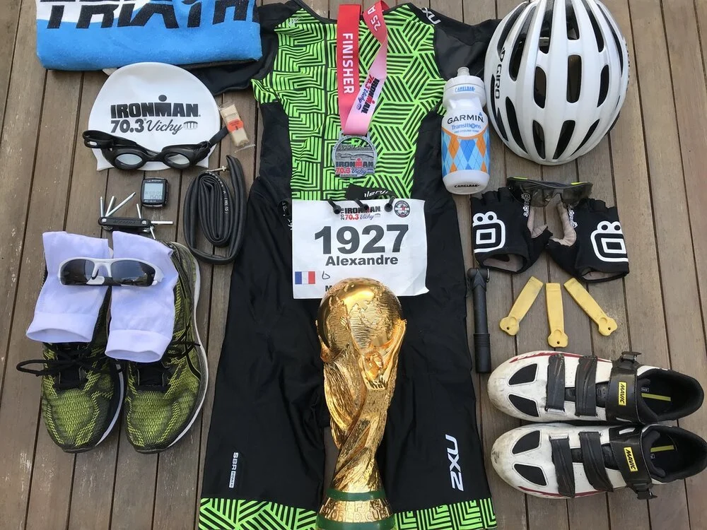Triathlon equipment including a bicycle helmet, swimming goggles, a race bib with the name Alexandre and the number 1927, a medal, running shoes, cycling shoes, a water bottle, a bicycle pump, a helmet, and various gear on a wooden surface.