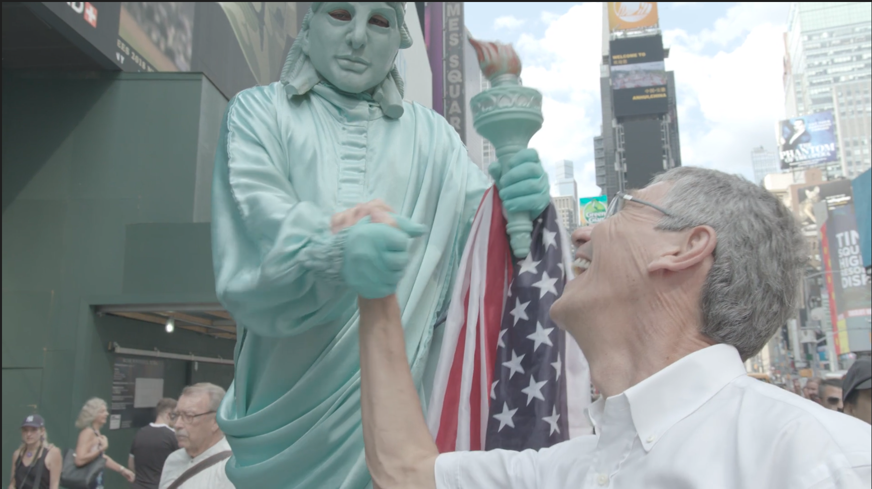 An older man with glasses smiling and shaking hands with a Statue of Liberty replica that is painted in a light blue color, holding an American flag in Times Square, New York City.