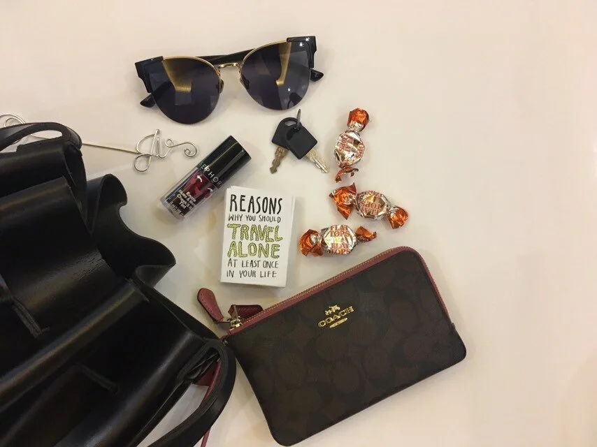 Sunglasses, a pair of earrings, a bottle of hand sanitizer, keys, a note that says "Reasons Why You Should Travel Alone At Least Once In Your Life," a bag of caramel candies, and a Coach wallet on a white surface.