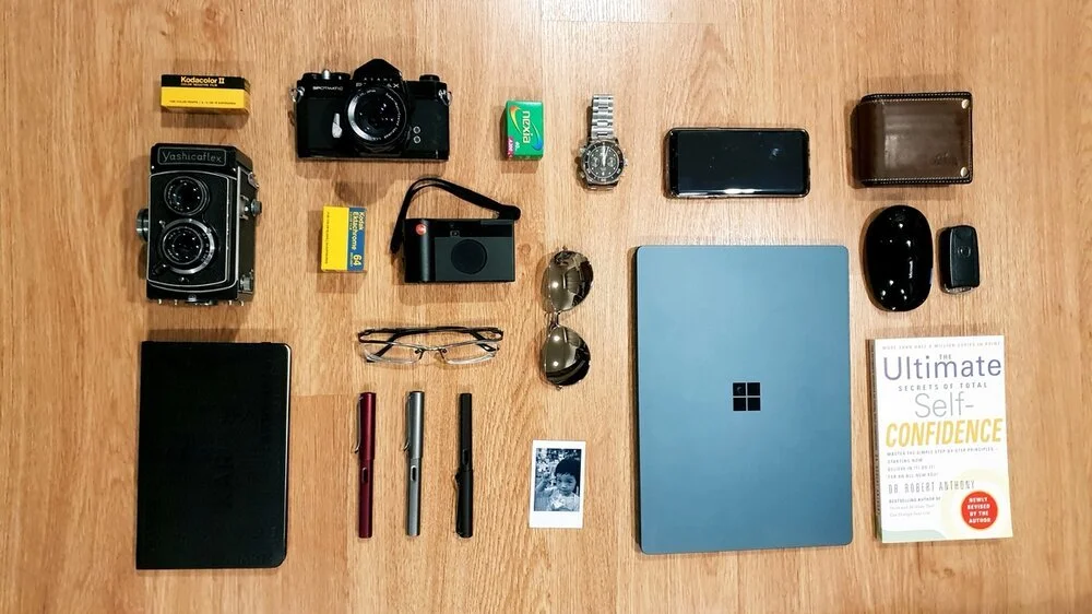 Flat lay of various items including a camera, film rolls, batteries, a small camera, sunglasses, a remote, a smartphone, a wallet, a wireless mouse, a key fob, a notebook, three pens, a photograph of a child, a laptop with a Windows logo, and a book 
