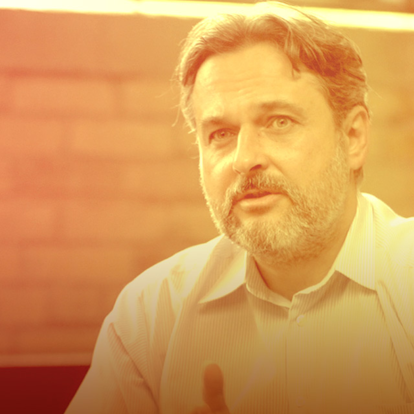A middle-aged man with brown hair and a beard, wearing a light-colored striped shirt, sitting indoors with warm lighting.