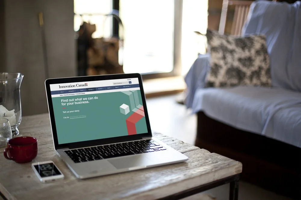 Laptop on a wooden table displaying the Innovation Canada website, with a glass of water, a small white device, and a red mug nearby, in a cozy room with a couch and pillows in the background.