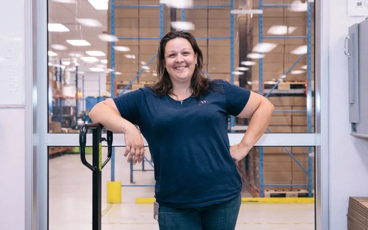 A woman with dark hair and a blue T-shirt smiling and standing in front of warehouse shelves.
