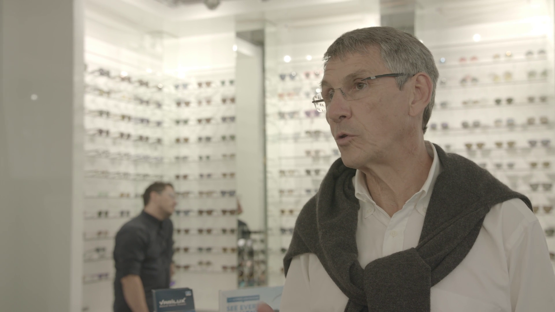 An older man with glasses and gray hair, wearing a white shirt and gray sweater, speaking in an eye glasses store with numerous eyeglasses displayed on the wall behind him. A younger man in black in the background at a counter.