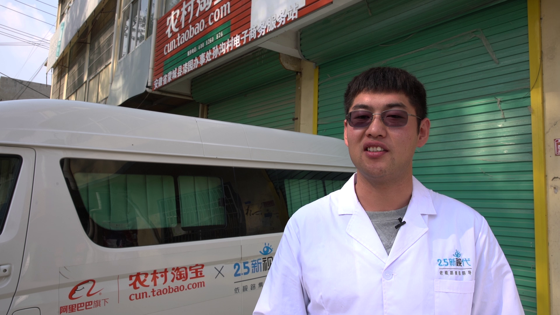 A man wearing sunglasses and a white coat standing outdoors next to a white vehicle with Chinese and English writing on it. Behind him are closed green shop shutters and a sign with Chinese characters and a URL for taobao.com.