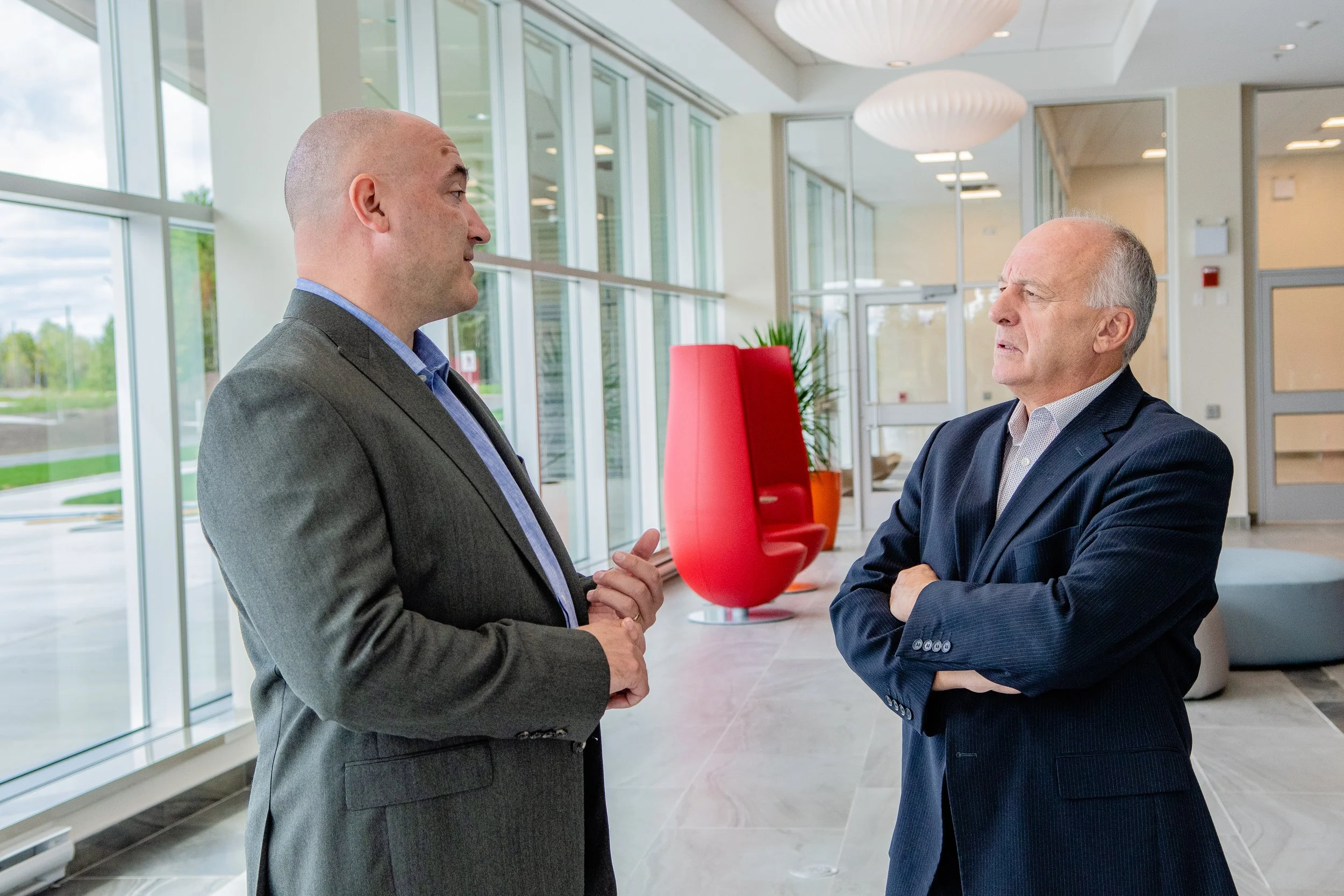 Two men in suits having a conversation in a modern lobby with floor-to-ceiling windows, a red chair, and potted plants.
