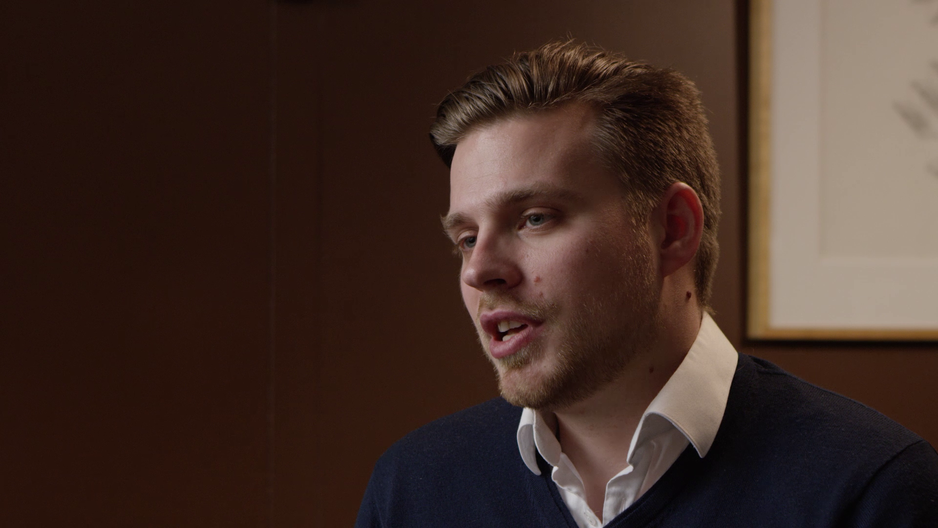 A young man with short, styled hair speaking in a close-up shot, wearing a white collared shirt and a dark sweater against a brown wall with a framed picture.