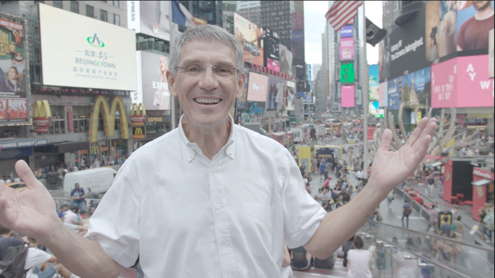 Man smiling and gesturing with open arms in Times Square, New York City, surrounded by billboards and a crowd of people.