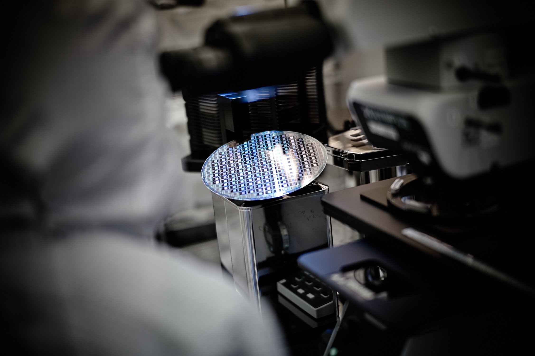 Close-up view of a semiconductor wafer with a reflective surface, placed on a workbench in a cleanroom or laboratory setting, surrounded by technical equipment.