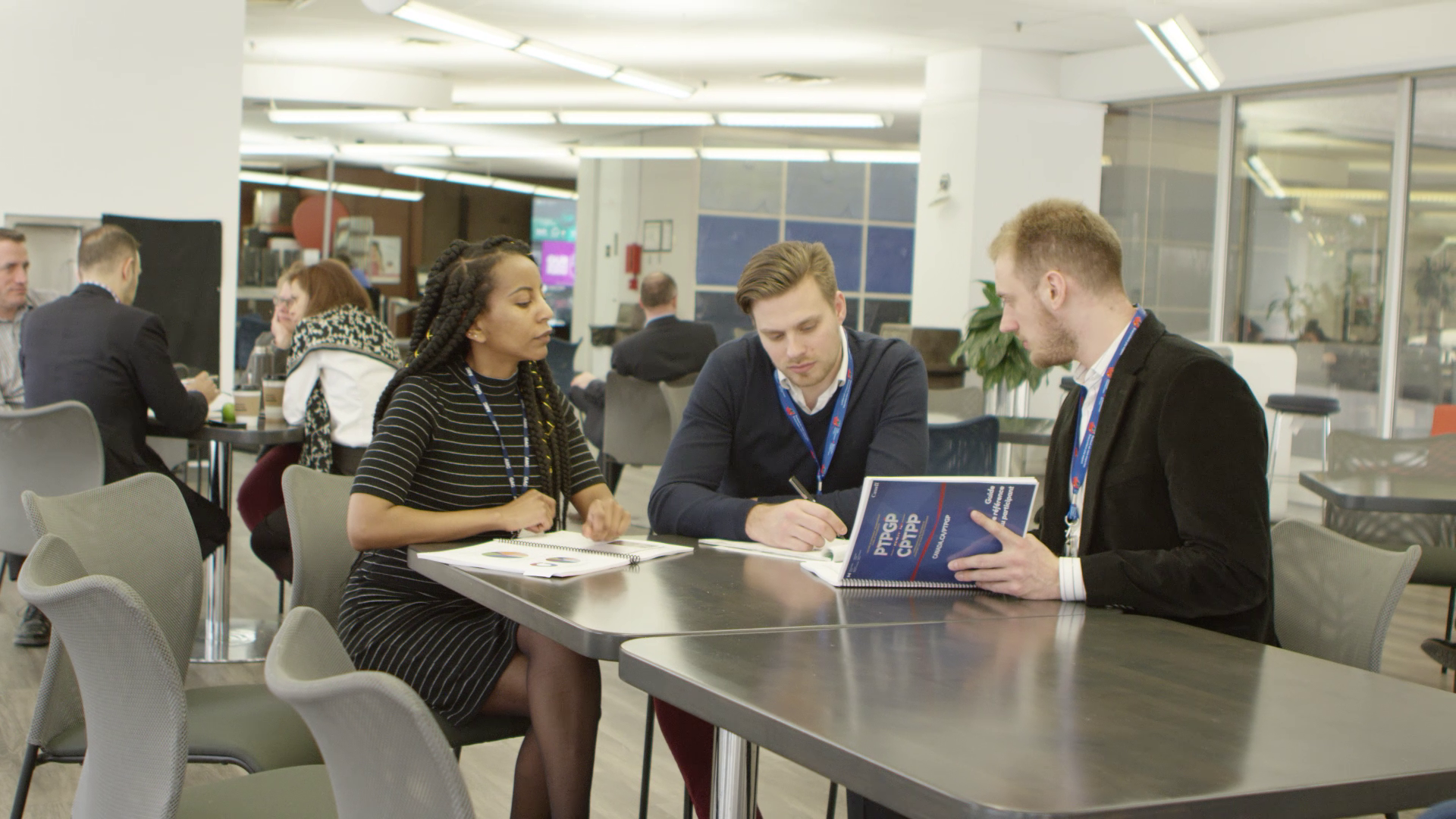 Three young adults sitting at a table in a modern conference or co-working space, engaged in a discussion or review of documents, with other people working in the background.