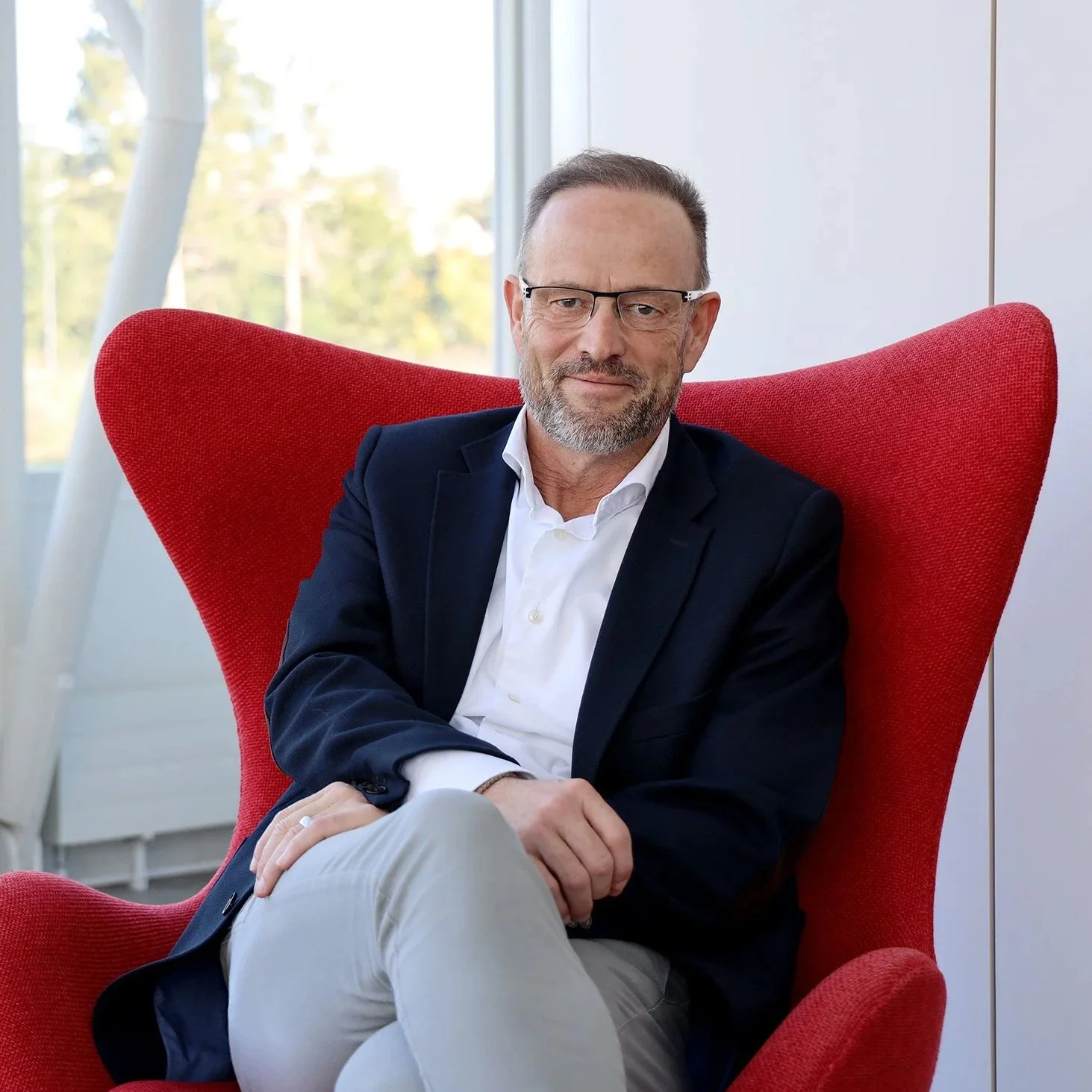 Ein Mann mit Brille, dunklem Anzug und weißem Hemd sitzt in einem roten Sessel vor einem Fenster mit Ausblick auf Bäume, lächelt leicht und wirkt entspannt.