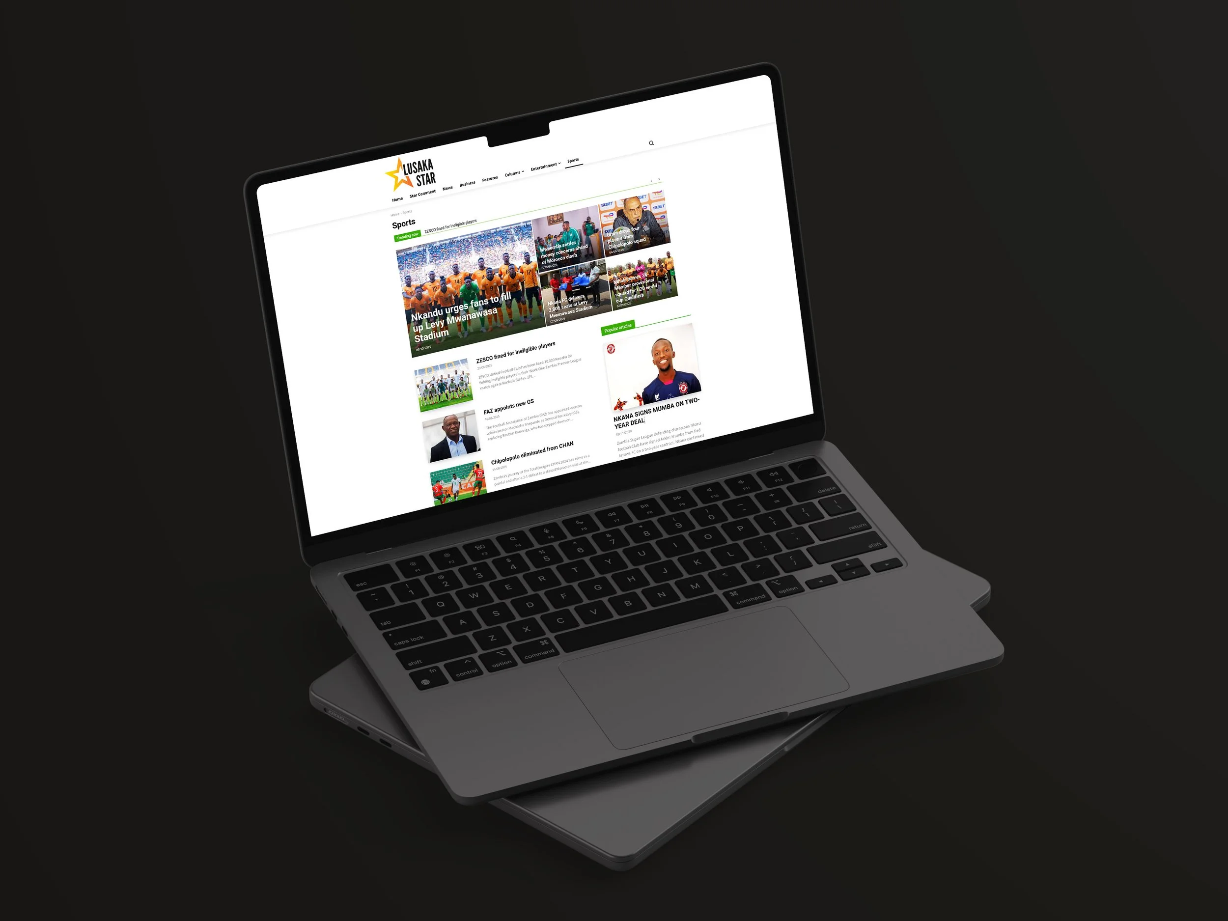 Ein Laptop auf einer schwarzen Oberfläche zeigt eine Webseite namens 'Lusaka Star' mit Sportnachrichten, darunter ein Gruppenfoto eines Fußballteams in orangefarbenen Trikots und weiterführende Artikel.