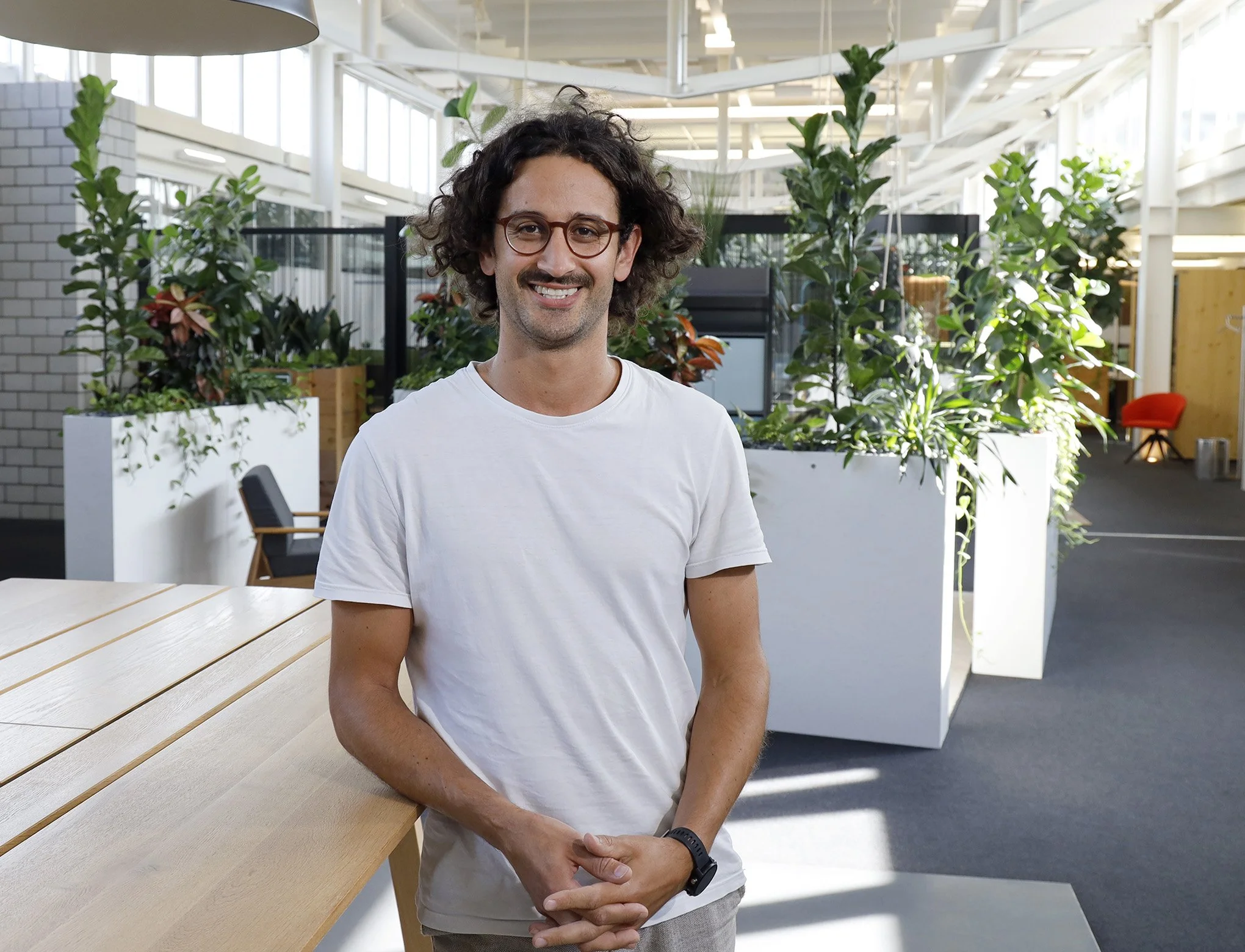 Junger Mann mit Brille und weißen T-Shirt in einem modernen Büro mit grünen Pflanzen und großen Fenstern.