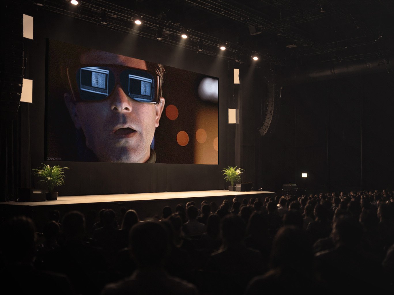 Menschenmenge sitzt in einem dunklen Eventraum, auf der Leinwand ist eine Person mit dunkler Computer-Brille zu sehen.